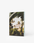 A hardcover book from Assouline, titled "Hotel Wailea," stands upright against a white background. The cover design features a tropical landscape with palm leaves and trees framing a distant mountain, reminiscent of the serene ambiance of the Maui boutique resort. The spine is light blue with subtle text, and the bottom of the cover displays "assouline.com".