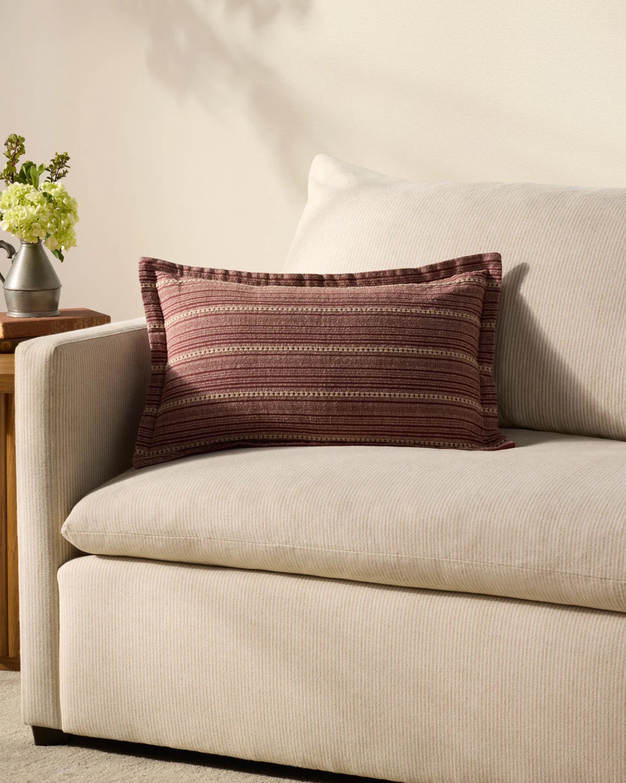 A cream sofa features the Loloi Rugs PCJ0023 Howie Wine Pillow 13x21, showcasing maroon and beige stripes. Next to it stands a wooden side table with an antique bronze finish and a flower vase against a light wall.
