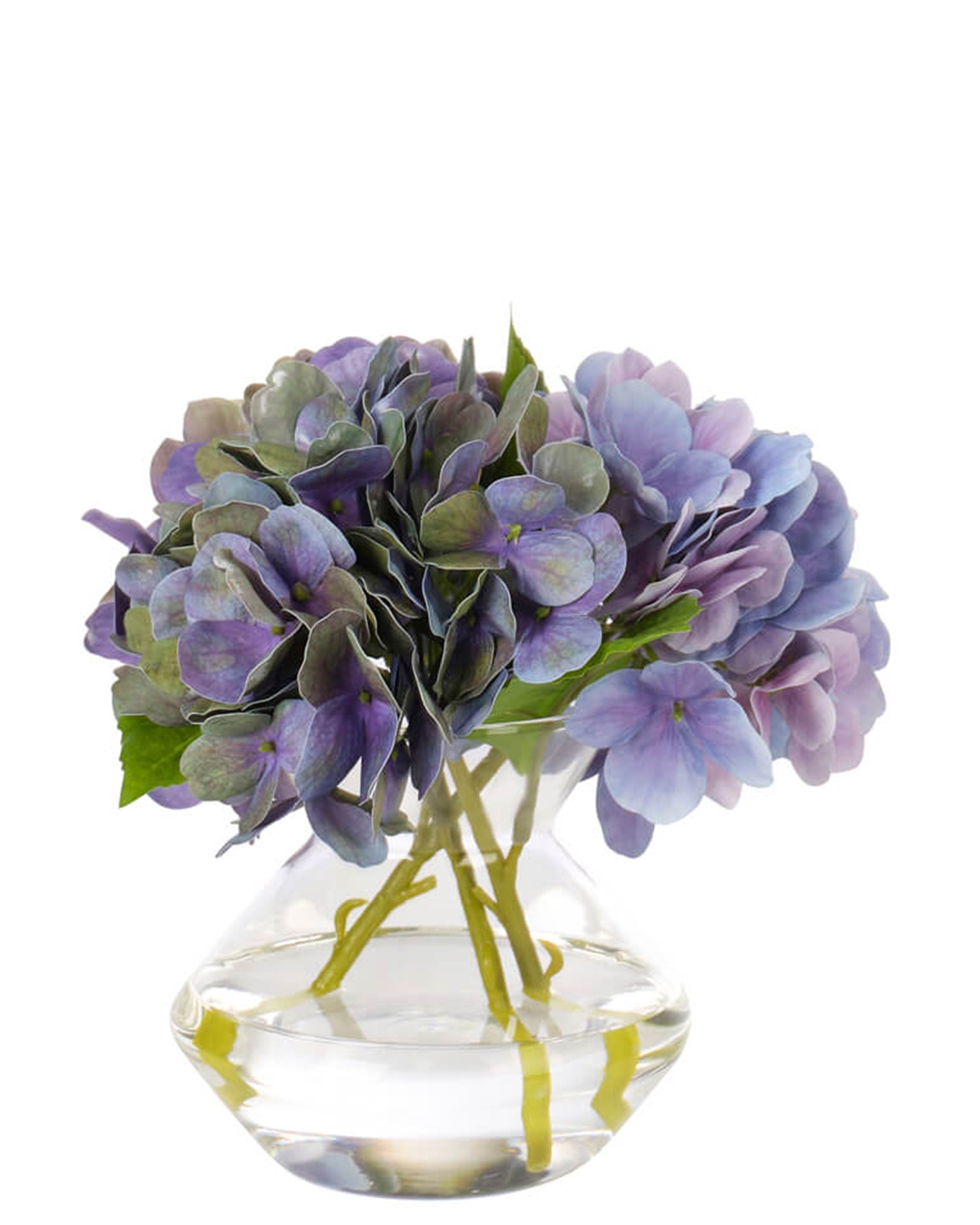 The Hydrangea Purple Blue by Natural Decorations Inc. features a clear glass vase with artificial purple and blue hydrangeas and green leaves, displayed against a white background.