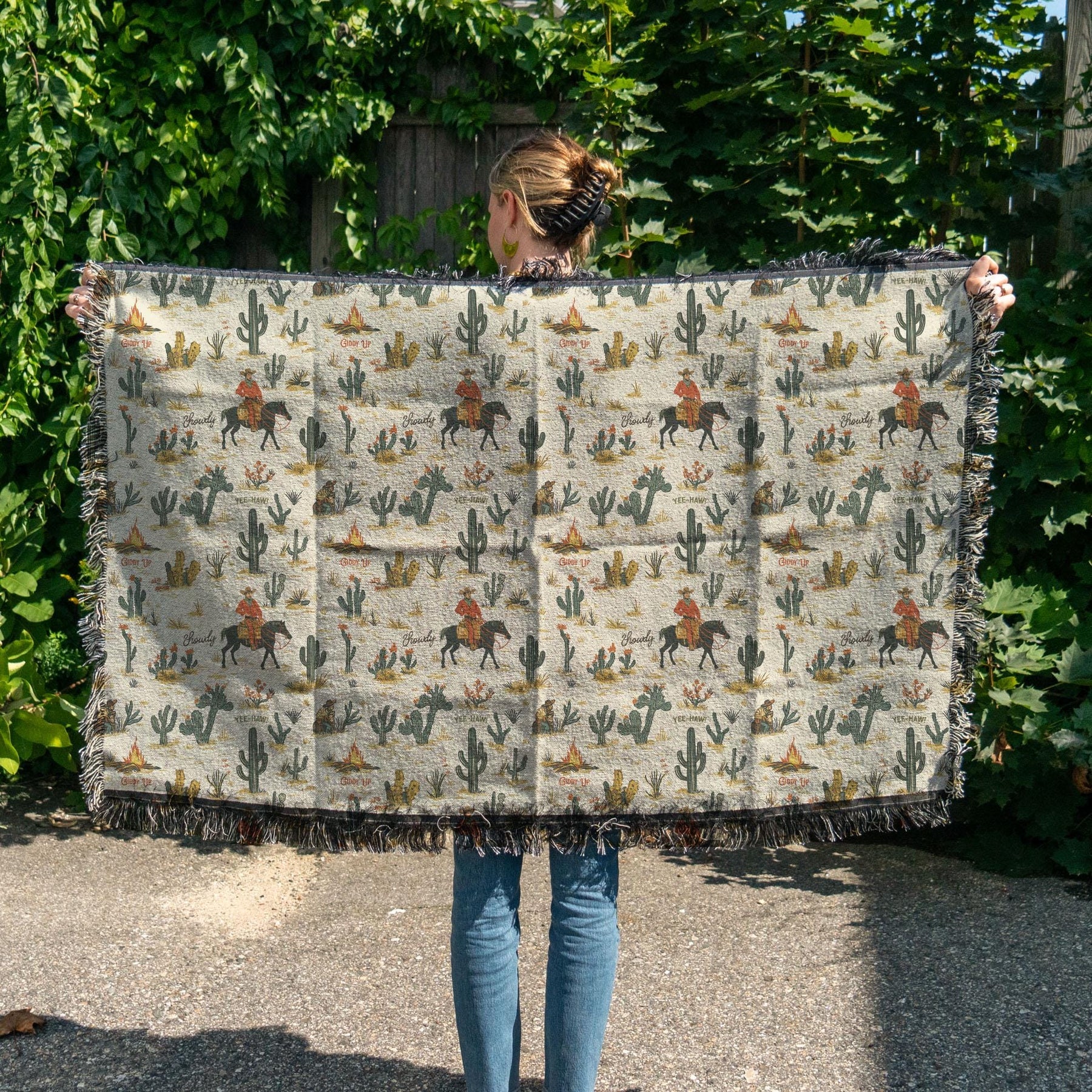 A person stands outdoors holding the Faire Western Blanket, which features cowboys on horses, cacti, and mountains in a desert-themed design. Lush green foliage appears in the background.