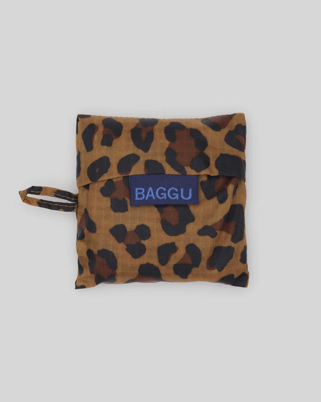 The Baby Baggu, a leopard print foldable bag by Baggu, crafted from recycled ripstop nylon with "BAGGU" on a blue tag, is set against a plain light grey background. This practical and stylish reusable shopping bag is machine washable for easy care.