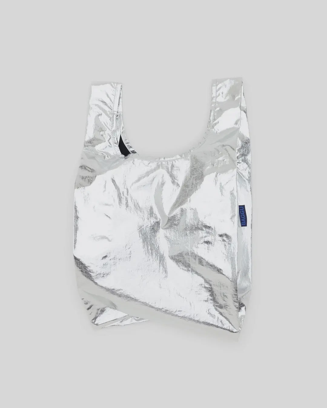 The Baby Baggu Metallic Silver tote by Baggu is a crinkled texture, recycled nylon bag with simple wide handles and a plain design. It rests against a light gray background, perfect for stylish shopping.