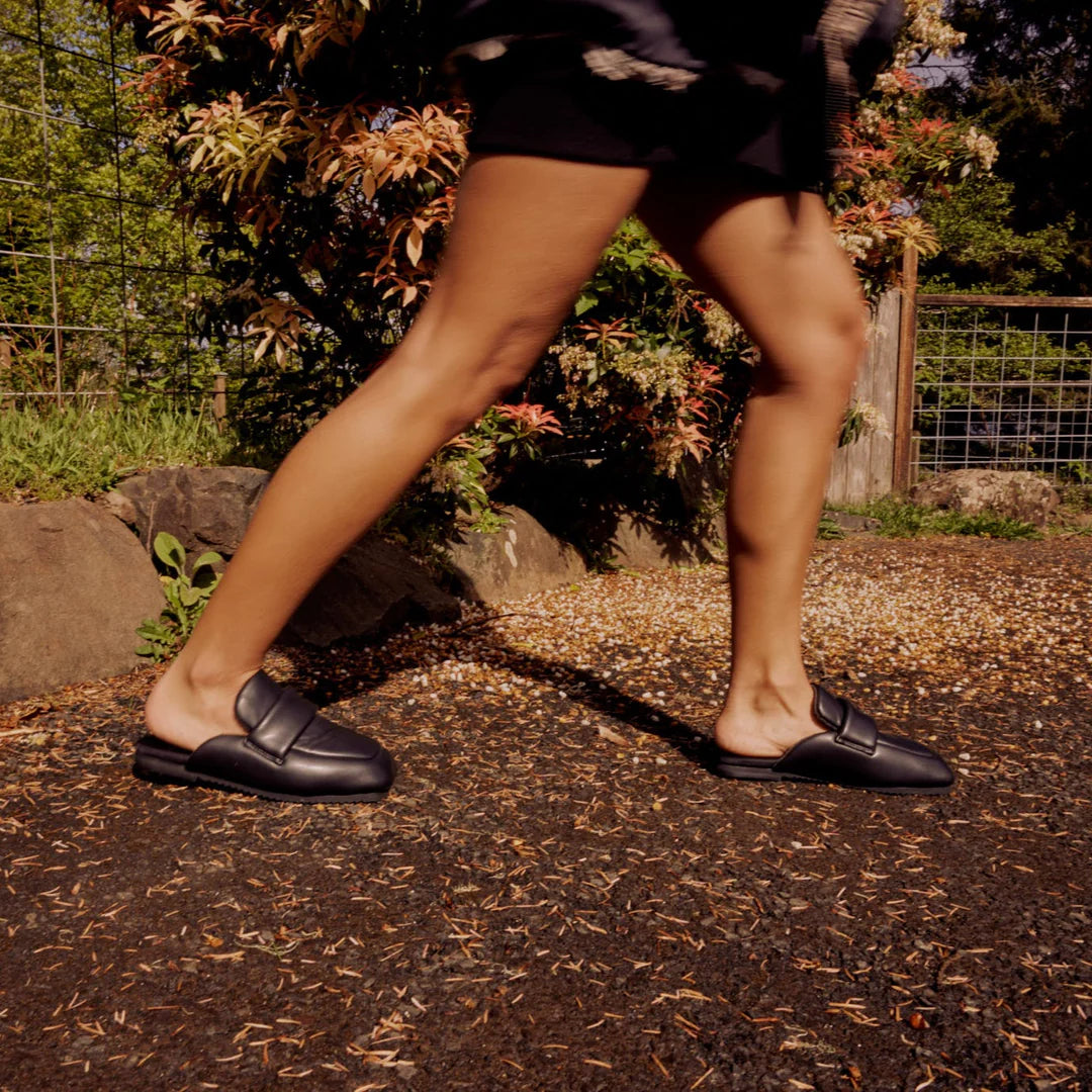 A person wearing Roam Puffy Loafers in black vegan leather and black shorts walks on a gravel path outdoors, with greenery and a fence behind them. The low-angle photo highlights their legs and the loafers.