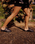 A person wearing Roam Puffy Loafers in black vegan leather and black shorts walks on a gravel path outdoors, with greenery and a fence behind them. The low-angle photo highlights their legs and the loafers.