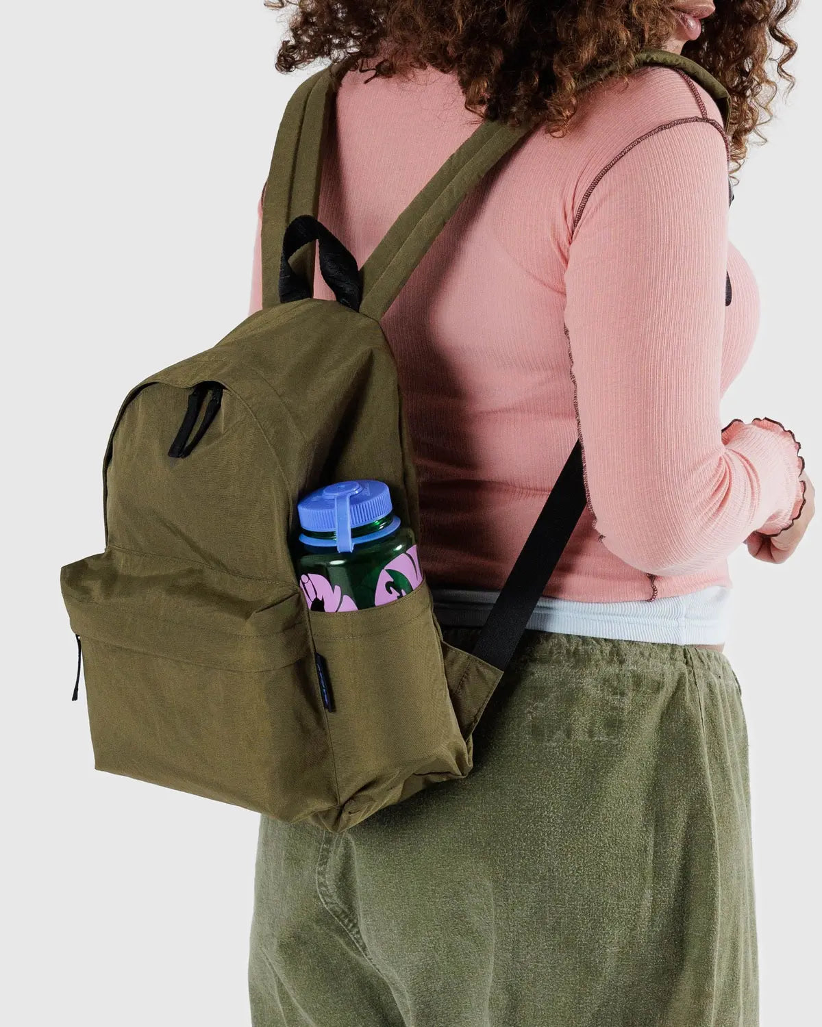 A person wearing a pink long-sleeve shirt and green pants carries a Baggu Nylon Backpack made from recycled nylon. The backpack, with adjustable straps and a side pocket, holds a colorful water bottle adorned with pink and green patterns.