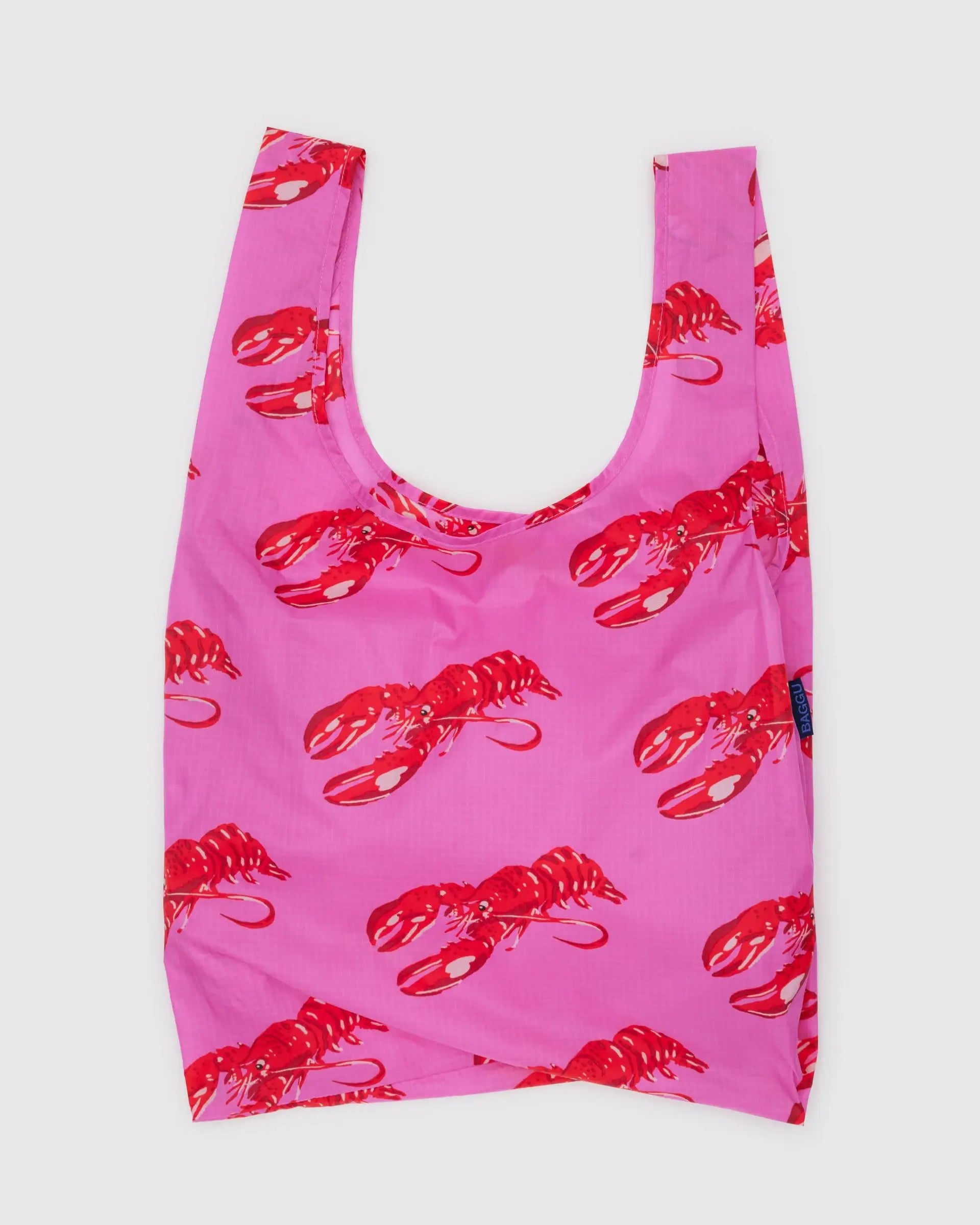 The Baggu Standard by Baggu is a reusable bag crafted from recycled ripstop nylon, featuring a pink backdrop with red lobster prints. It offers wide handles, a slightly glossy finish, and is machine washable for daily use.