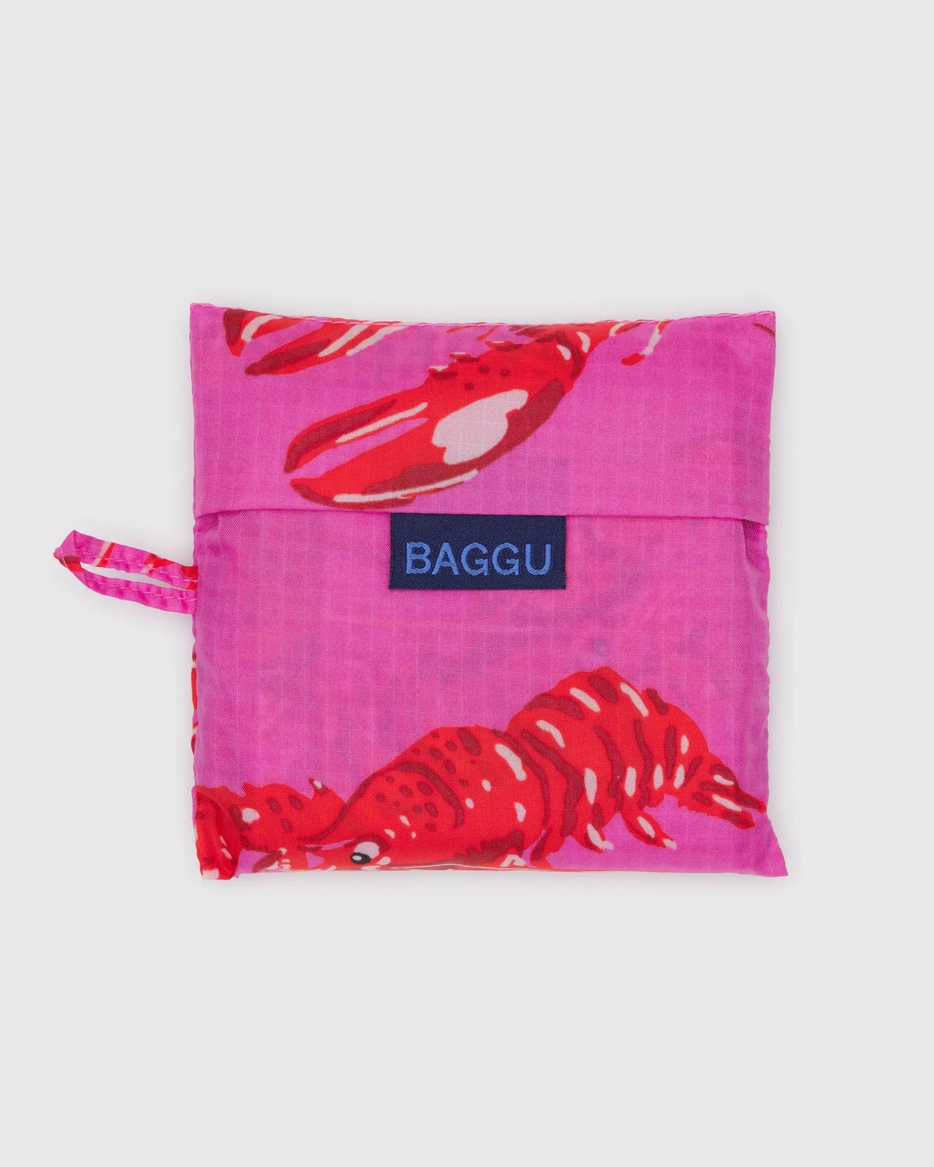 The Baggu Standard by Baggu is a folded, reusable shopping bag made from recycled ripstop nylon with a bright pink background, red lobster print, a small left-side handle loop, and a visible BAGGU logo label on the front.