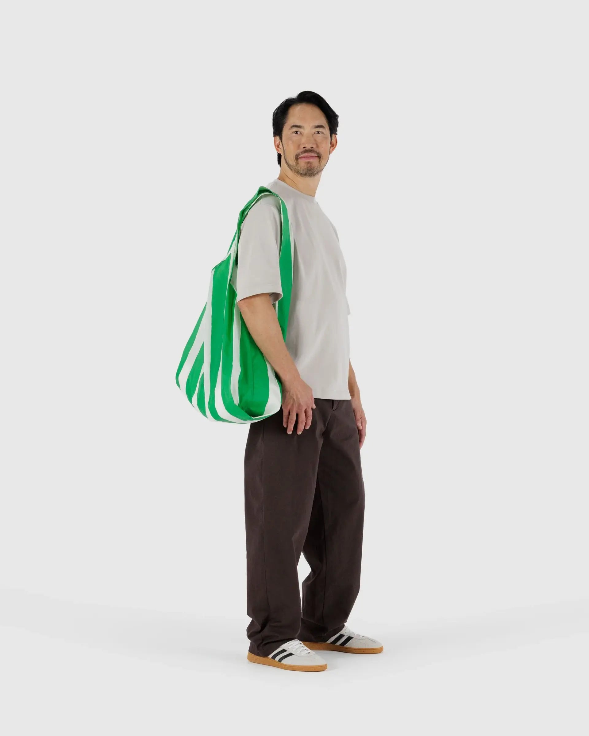 A man stands against a plain background, wearing a white T-shirt, dark brown pants, white sneakers, and carrying the large green and white striped Big Baggu recycled ripstop nylon shopping bag by Baggu over his shoulder.