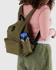 A person wearing a pink long-sleeve shirt and green pants carries a Baggu Nylon Backpack made from recycled nylon. The backpack, with adjustable straps and a side pocket, holds a colorful water bottle adorned with pink and green patterns.