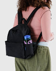 Someone with curly hair is dressed in a pink long-sleeve top and olive green pants, carrying Baggu's Nylon Backpack in black with adjustable straps. A colorful water bottle with a blue cap and pink design is tucked into the side pocket of the recycled nylon bag.