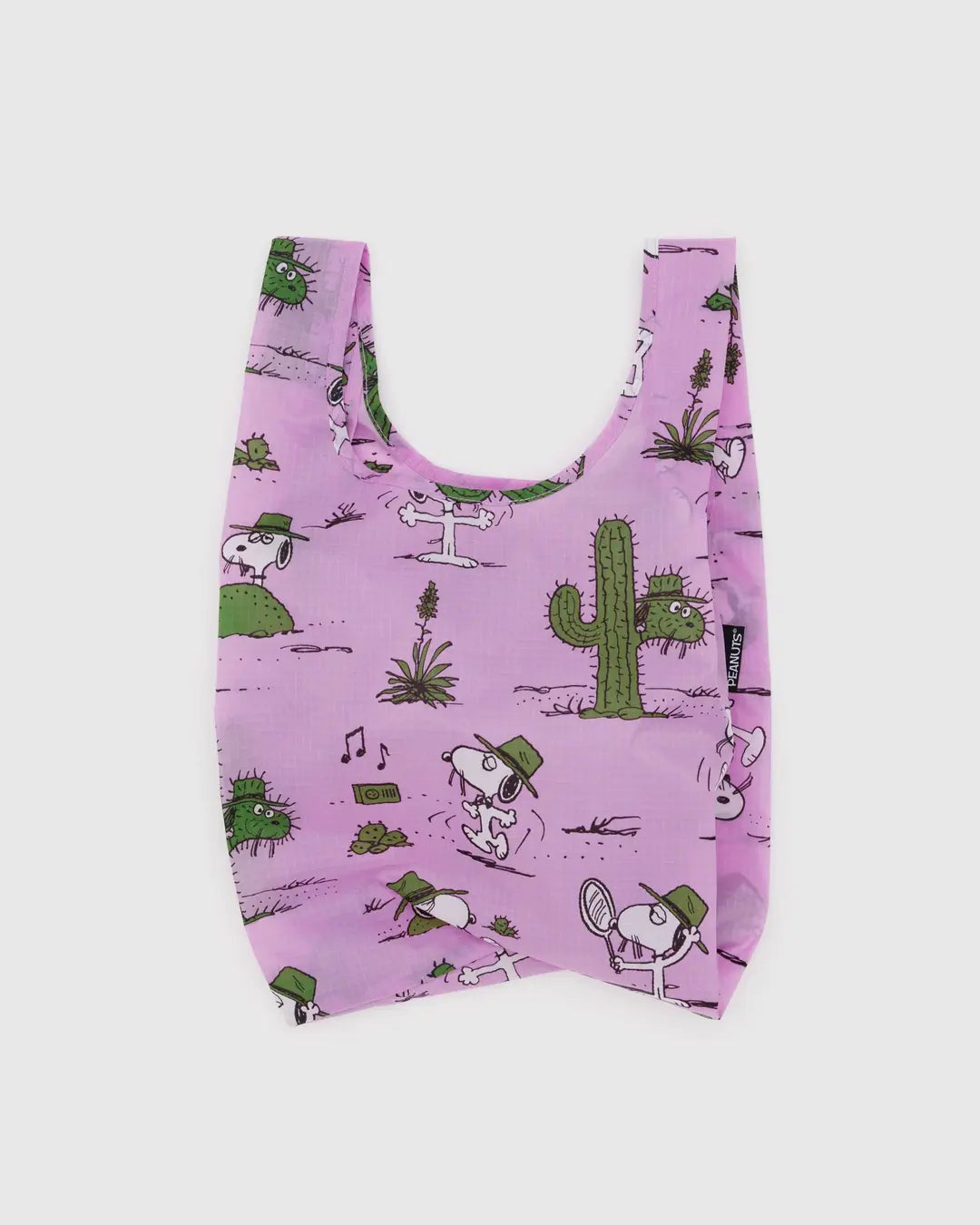 The Baby Baggu by Baggu is a reusable shopping bag made from recycled ripstop nylon, featuring a pink background with cartoon dogs in green hats and a playful desert-themed pattern of cacti, musical notes, and plants. It's also machine washable.