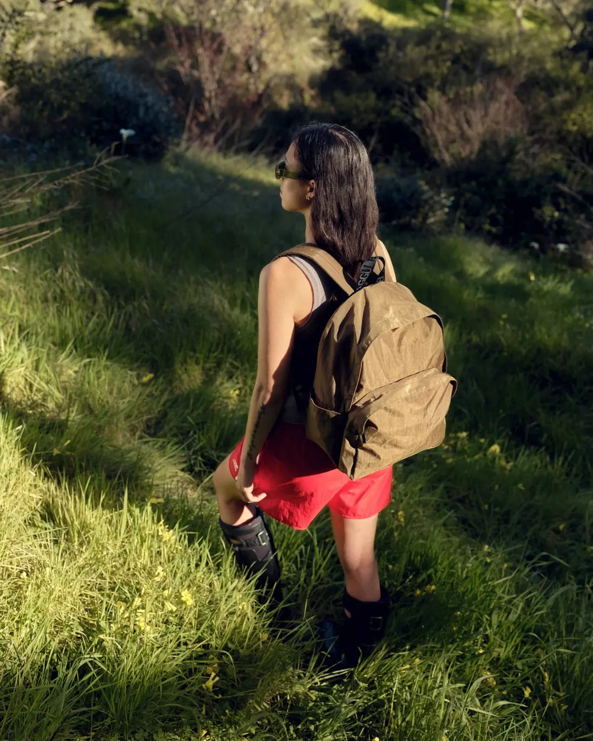 A person with dark hair stands on a grassy path in a sunlit forest, wearing a black top, red shorts, boots, and sunglasses. They carry an eco-friendly brown Baggu Nylon Backpack made from recycled nylon with adjustable straps while gazing into the distance.