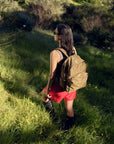 A person with dark hair stands on a grassy path in a sunlit forest, wearing a black top, red shorts, boots, and sunglasses. They carry an eco-friendly brown Baggu Nylon Backpack made from recycled nylon with adjustable straps while gazing into the distance.