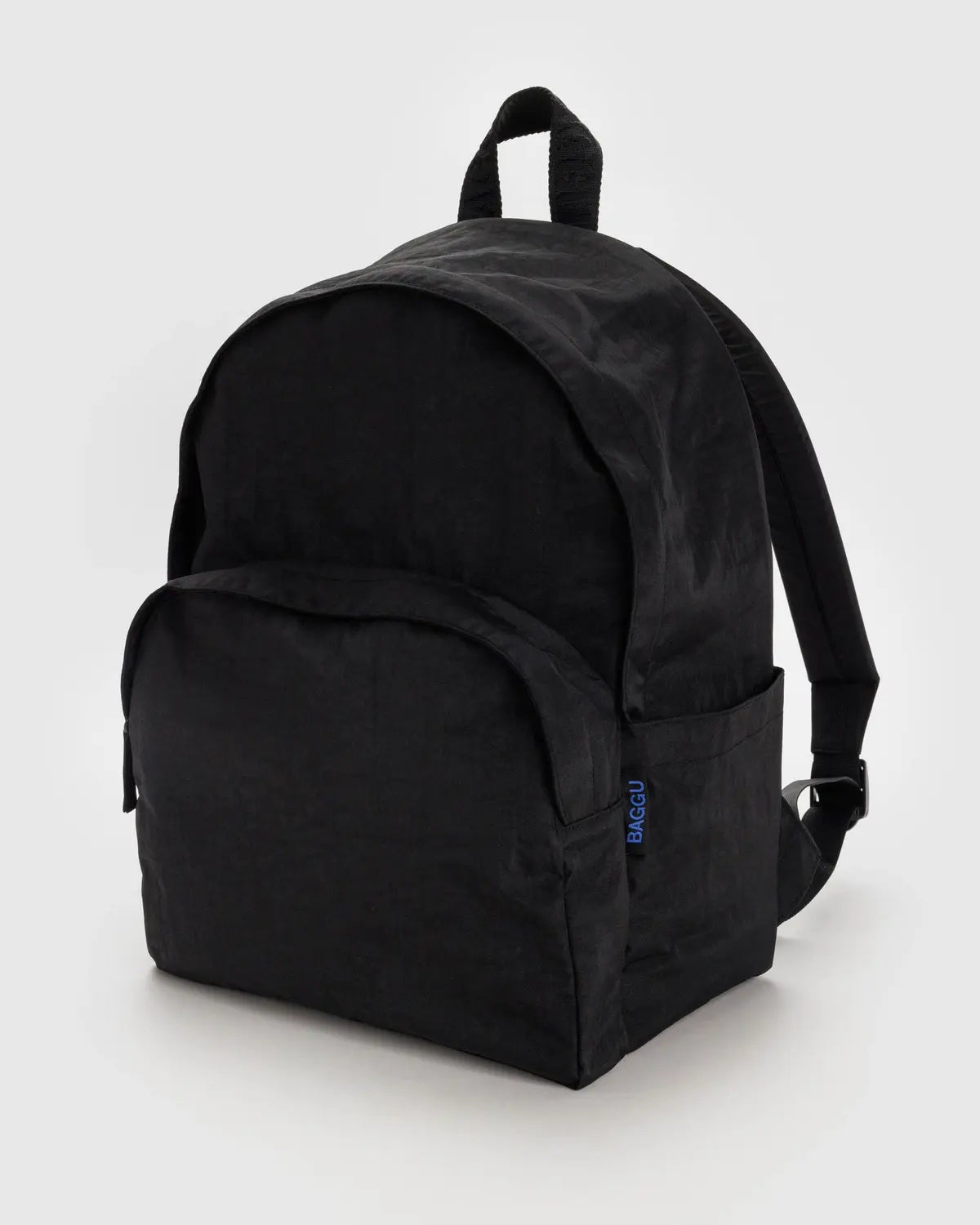 The Baggu Nylon Backpack, made from recycled black nylon, is shown against a plain background. It has a rounded top, front pocket with a small blue tag, top handle, adjustable shoulder straps, and includes a laptop sleeve.