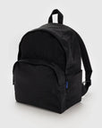 The Baggu Nylon Backpack, made from recycled black nylon, is shown against a plain background. It has a rounded top, front pocket with a small blue tag, top handle, adjustable shoulder straps, and includes a laptop sleeve.