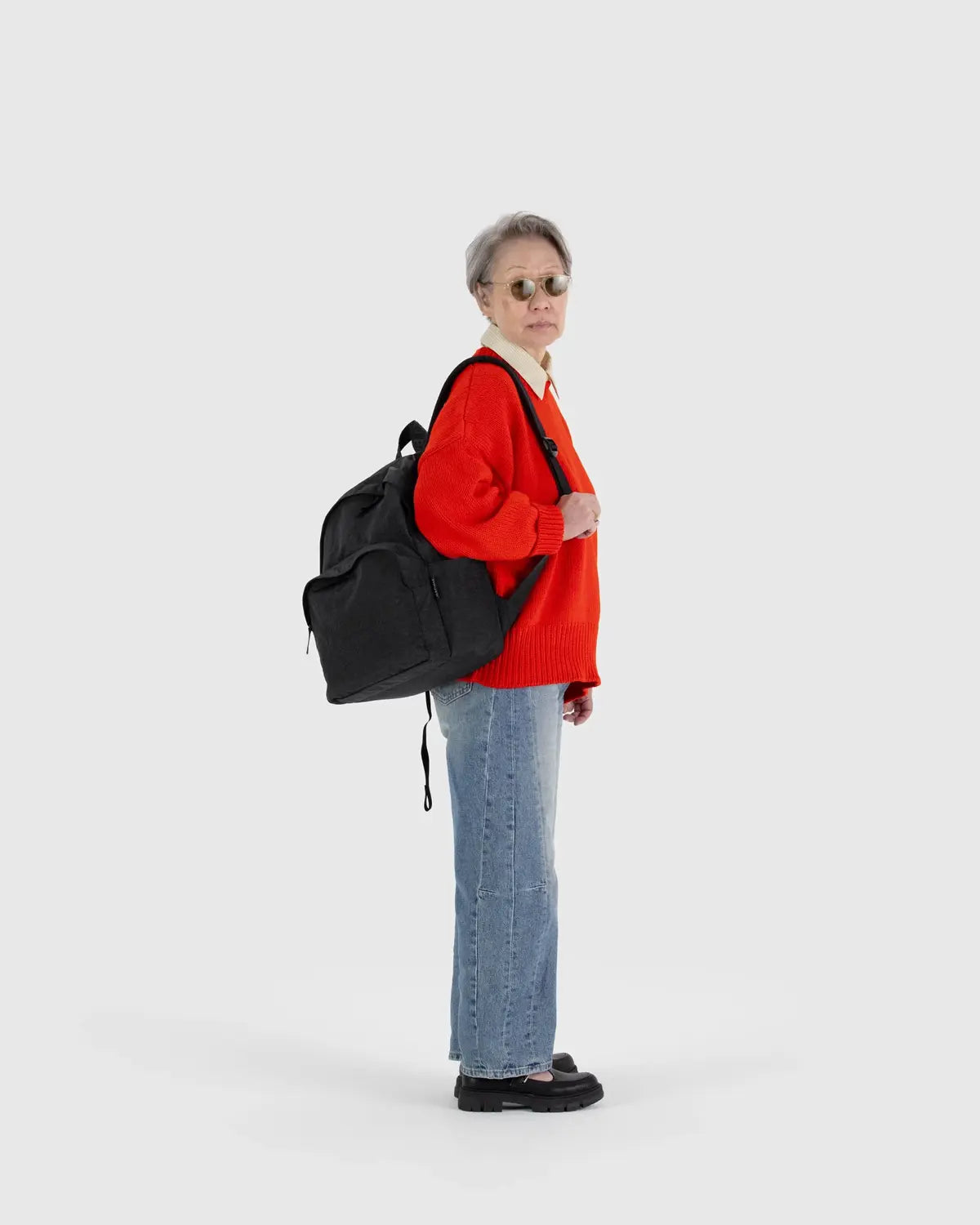 An older adult with short gray hair wears sunglasses, a red sweater, blue jeans, and black shoes. They carry Baggu’s Nylon Backpack in sleek black, made from recycled nylon with adjustable straps, and stand sideways against a plain gray background.