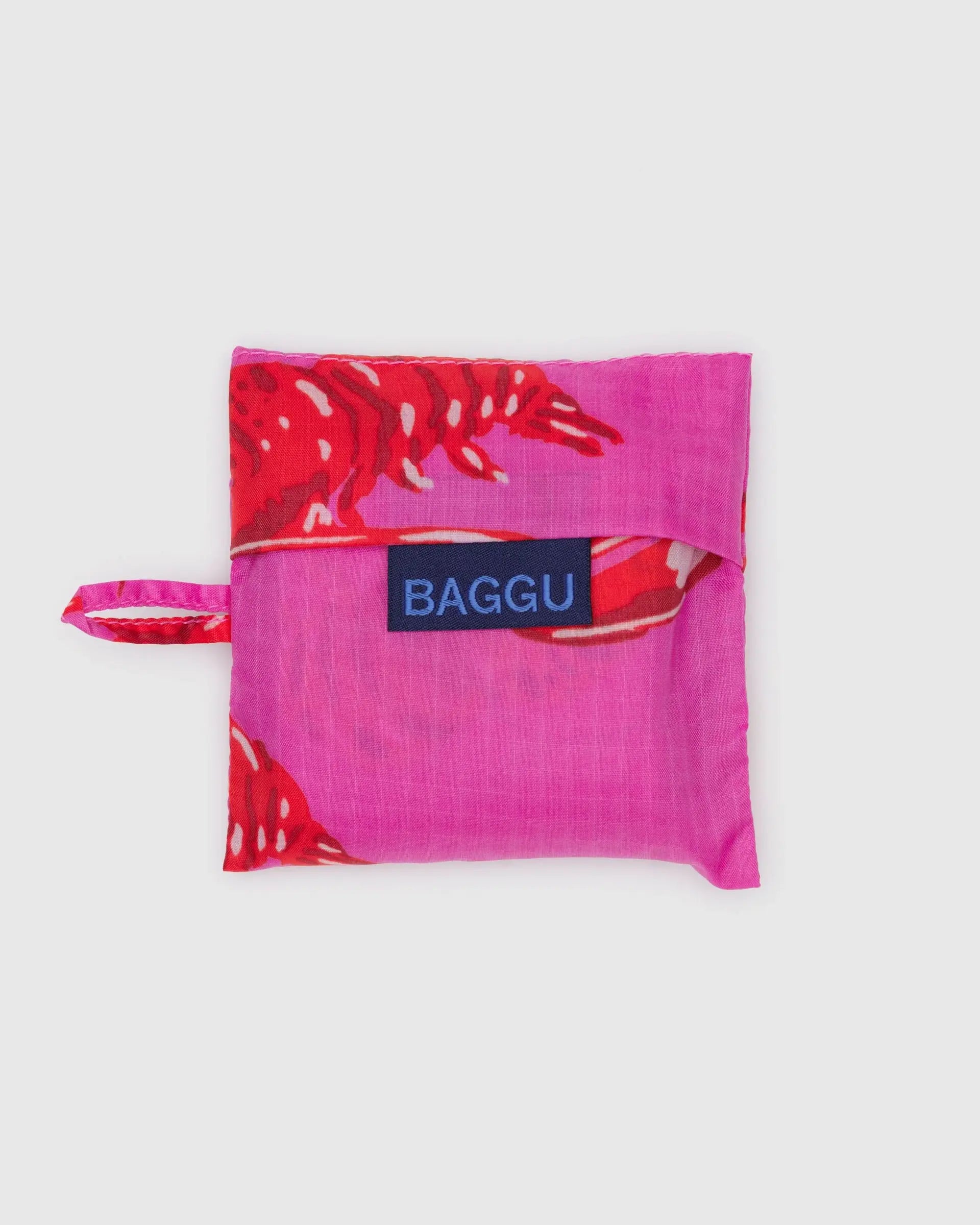 A folded pink Baby Baggu reusable shopping bag by Baggu, featuring a red lobster print, packed into its pouch with a small loop and blue Baggu label on the front, shown against a plain white background.