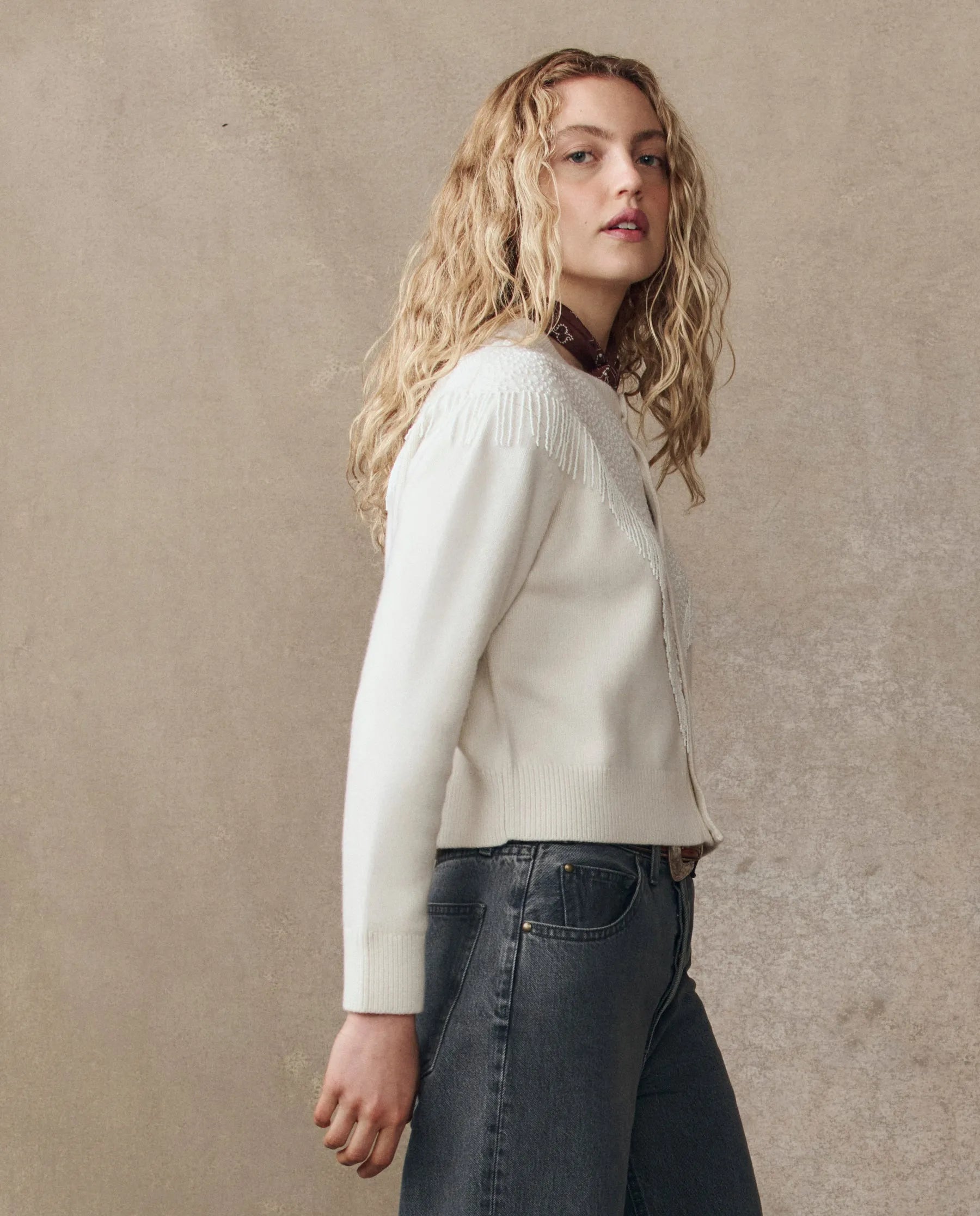 A woman with long, wavy blonde hair stands sideways in The Great Inc. Beaded Fringe Cardigan and high-waisted gray jeans against a plain beige background.