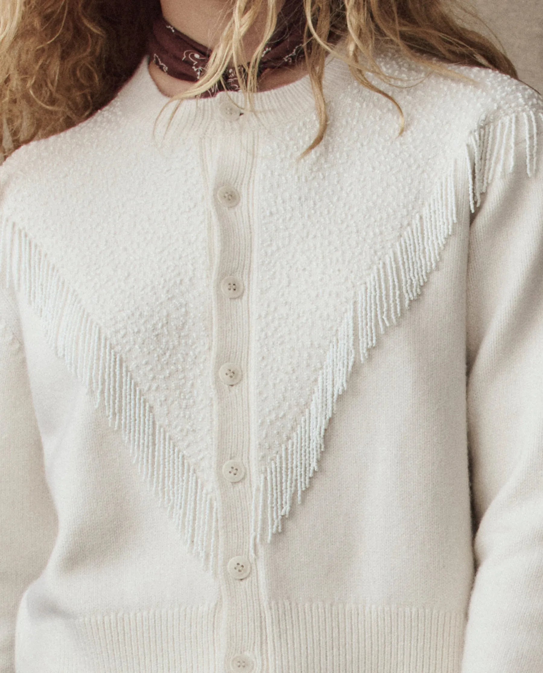 A person with wavy blonde hair wears The Great Inc. Beaded Fringe Cardigan, a cream knit with textured chevron patterns and beaded fringe, buttoned down the front, styled with a maroon bandana around their neck.