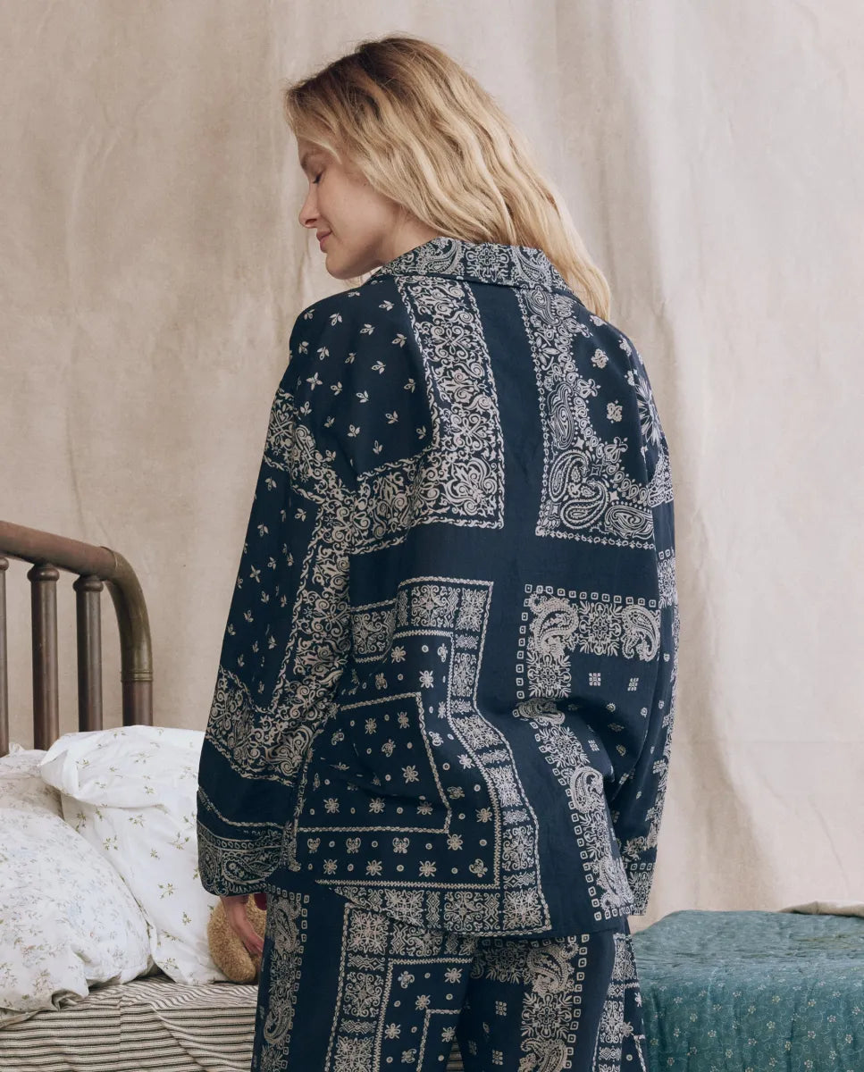 A blonde woman stands sideways on a bed, wearing The Great Inc.’s The Pajama Shirt in navy with a white bandana print. Beige curtains and a metal bed frame with white and green bedding are in the background.