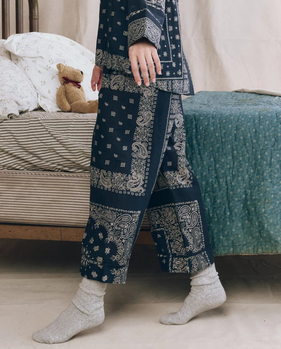 Someone wearing The Great Inc. Easy Sleep Pant in blue bandana patchwork print with gray socks stands by a bed featuring green and white bedding, a teddy bear resting on the pillows.