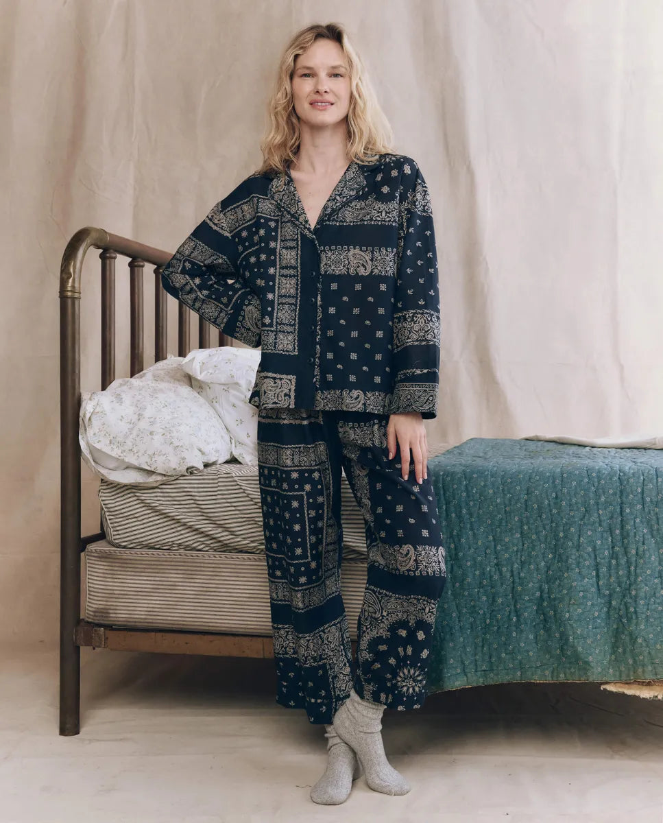 A person stands by a vintage-style metal bed, wearing The Great Inc. Easy Sleep Pant in high waist bandana patchwork print and light gray socks, one hand on the bed and a soft smile. The bedding features layered fabrics.