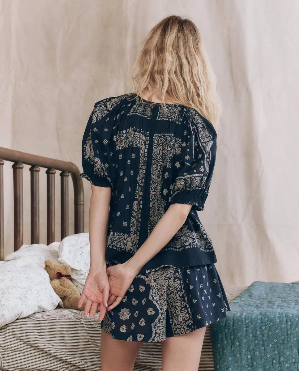 A woman with wavy blonde hair stands with her back to the camera, wearing The Sleep Short by The Great Inc.—a navy blue, bandana patchwork print short—in a bedroom with floral bedding and a teddy bear.