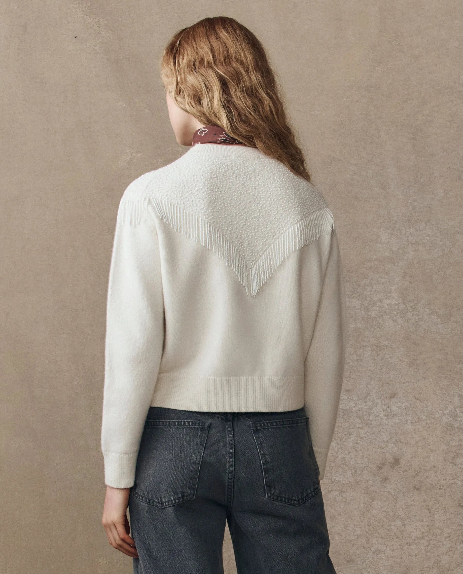 A person with wavy, light brown hair faces away, wearing The Beaded Fringe Cardigan by The Great Inc.—a cream-hued cardigan with a textured V-shaped pattern and beaded fringe on the back—paired with high-waisted, dark gray jeans. Plain beige background.