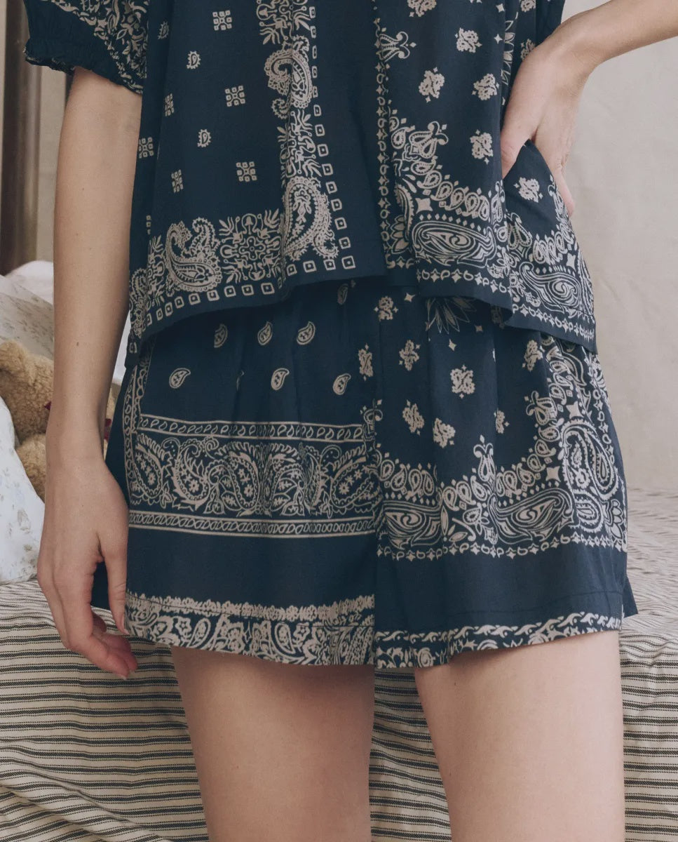 A person models The Great Inc.’s The Sleep Short—a matching navy blue set with a white paisley floral bandana patchwork print—posing with one hand on their hip against a neutral background.