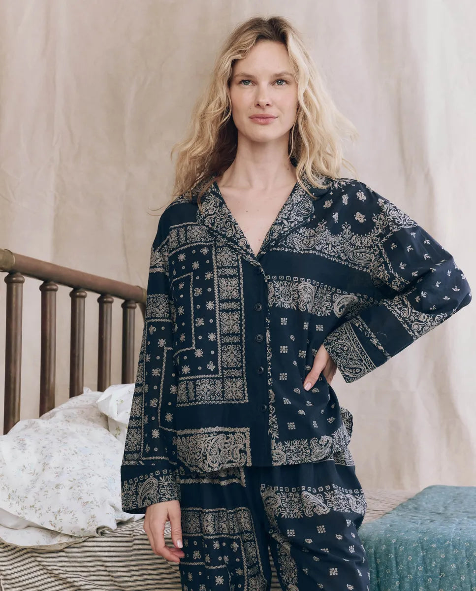 A woman with wavy blonde hair stands indoors by a wooden bed, wearing The Pajama Shirt by The Great Inc.—a vintage-inspired, navy blue and white patterned women's sleepwear top—with one hand on her hip and a neutral expression.