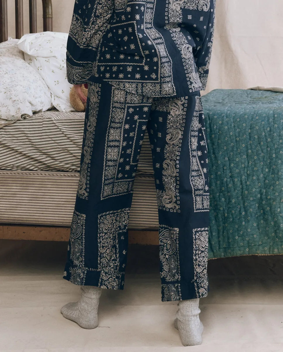A person wearing The Great Inc. Easy Sleep Pant in high waist navy blue cotton with a white bandana patchwork print stands by a bed with striped and floral bedding, light gray socks completing the cozy look.