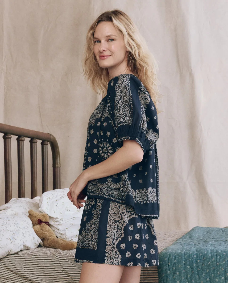 A woman with wavy blonde hair stands by a bed in The Sleep Short pajama set by The Great Inc., featuring navy blue patterned elastic waist shorts and a short-sleeve top. A teddy bear is on the bed and a green bench is nearby.