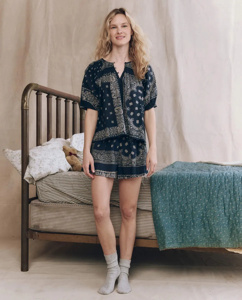A woman with wavy blonde hair smiles in front of a vintage metal bed, wearing The Great Inc.'s The Sleep Short in navy Bandana Patchwork Print with an elastic waist and gray socks. The bed has blue and white bedding and a teddy bear on the pillows.