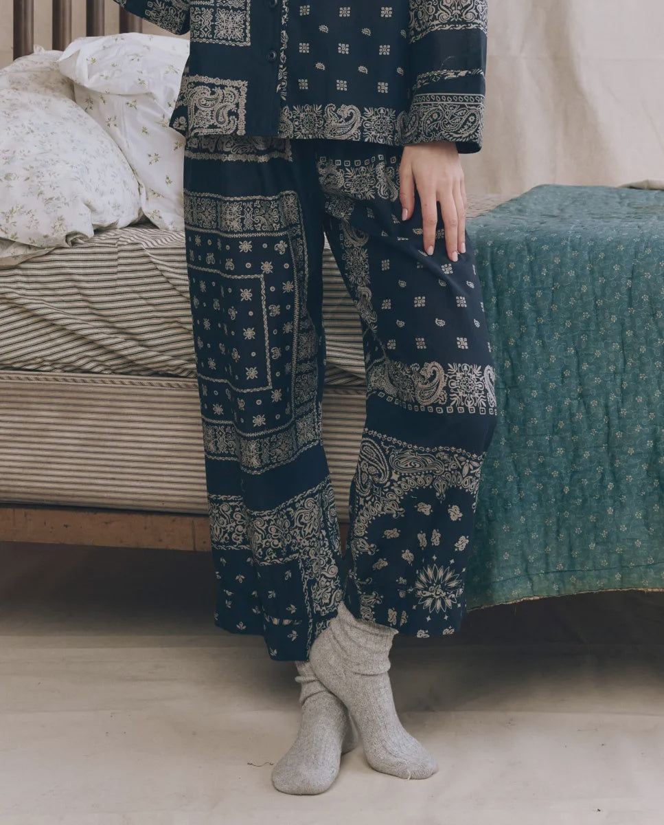 A person wearing The Easy Sleep Pant by The Great Inc. and gray socks stands by a bed with striped sheets and a green quilt, one hand resting on their thigh.