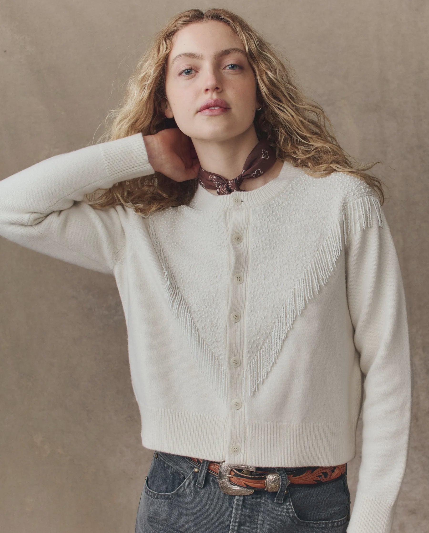 A woman with long curly blonde hair wears The Beaded Fringe Cardigan by The Great Inc., featuring fringe accents, paired with a brown patterned bandana, gray jeans, and a brown tooled leather belt against a neutral background.