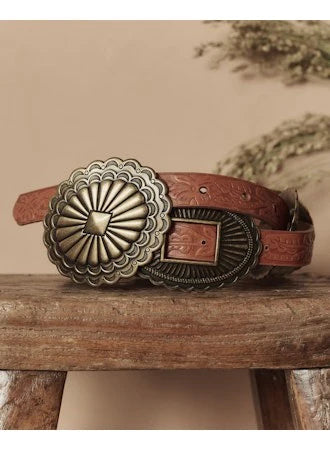The Leather Concho Belt by The Great Inc., featuring tooled black leather, embossed patterns, and two large ornate brass buckles, is displayed on a rustic wooden stool against a beige background.