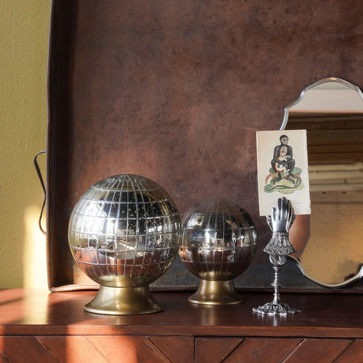 Two Blue Ocean Traders Wunderkammer Orbs on brass stands sit on wood beside a mirror held by a silver hand, which displays a vintage portrait card. A brown metal backdrop frames this distinctive vintage scene.