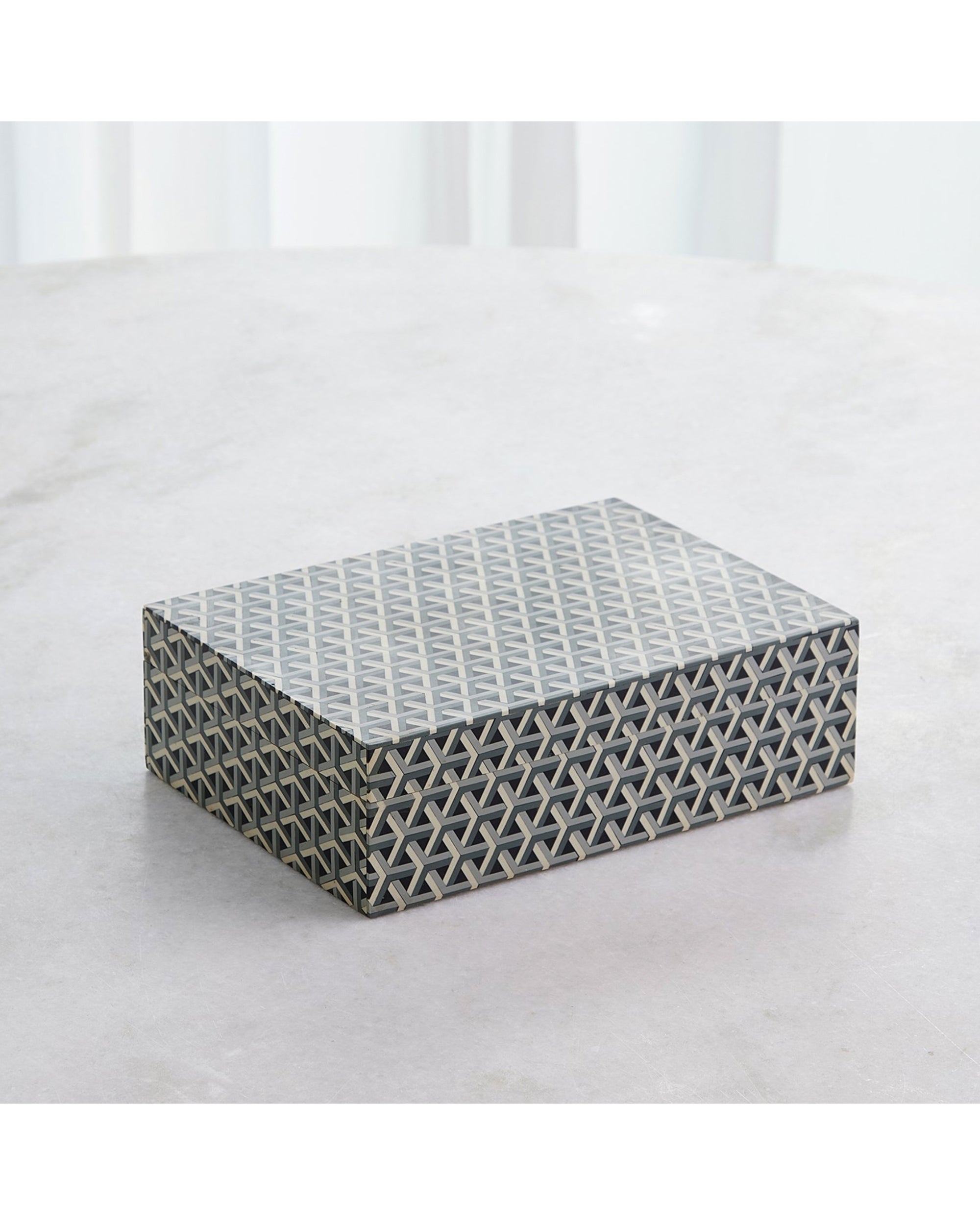 The 9.94040 Interwoven Box MD by Global Views is a rectangular decorative box featuring a black, white, and gray geometric star pattern, shown on a light surface with sheer white curtains in the background.