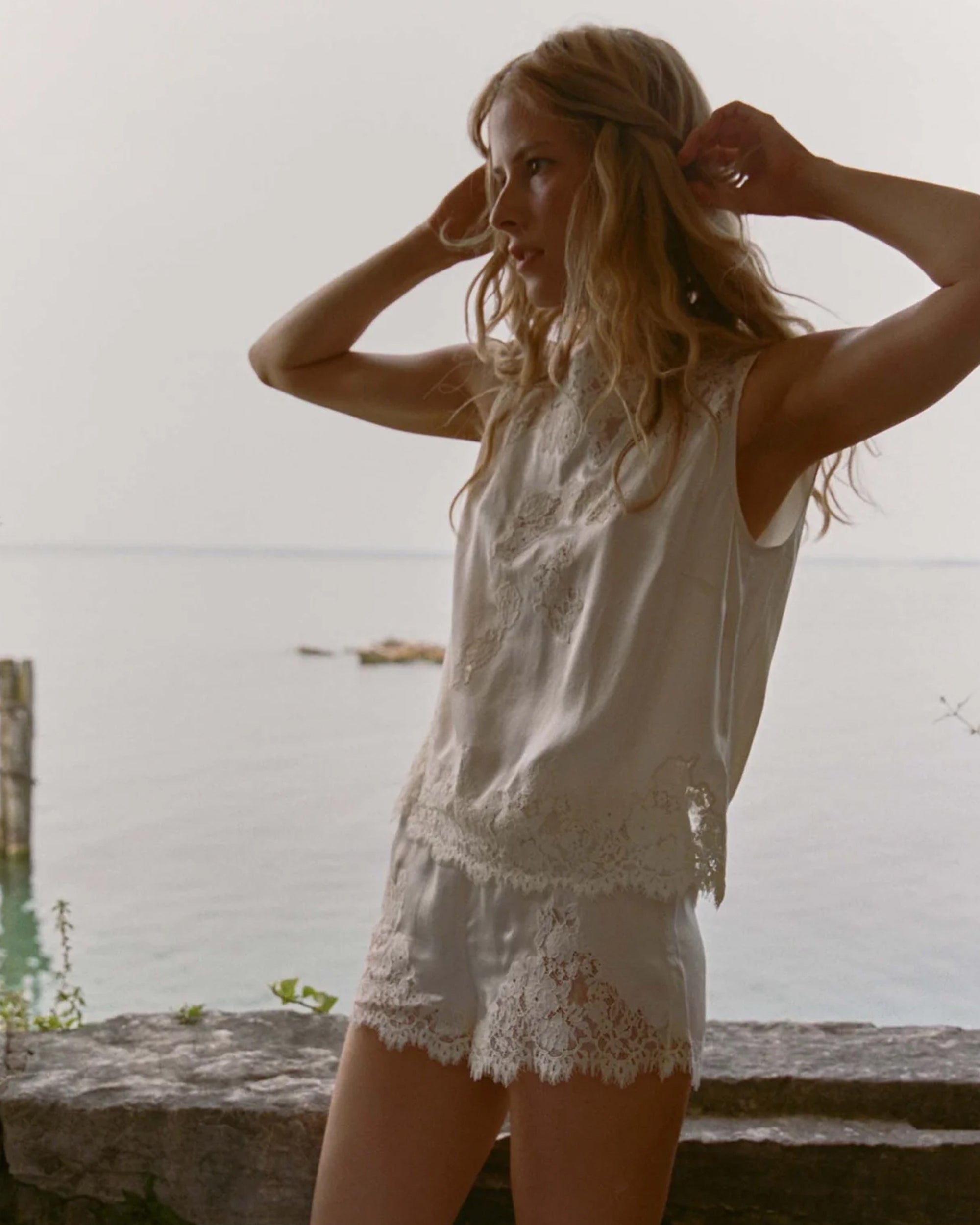 A woman wearing the DOEN Iona Short and a sleeveless floral lace white top stands outdoors by stonework with water and a hazy sky behind her, adjusting her hair and gazing to the side.