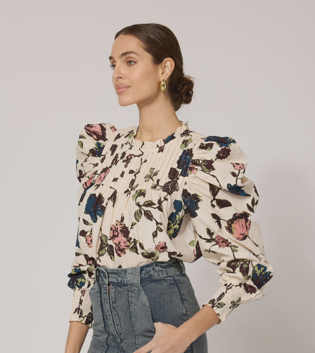 A person with long hair tied back is wearing the Irina Blouse from Cleobella, made from GOTS certified organic cotton and featuring ruffled shoulders, along with blue jeans. They are standing in front of a plain background, looking to the right, showcasing gold hoop earrings and a calm expression in their vintage-inspired look.