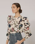 A person with long hair tied back is wearing the Irina Blouse from Cleobella, made from GOTS certified organic cotton and featuring ruffled shoulders, along with blue jeans. They are standing in front of a plain background, looking to the right, showcasing gold hoop earrings and a calm expression in their vintage-inspired look.