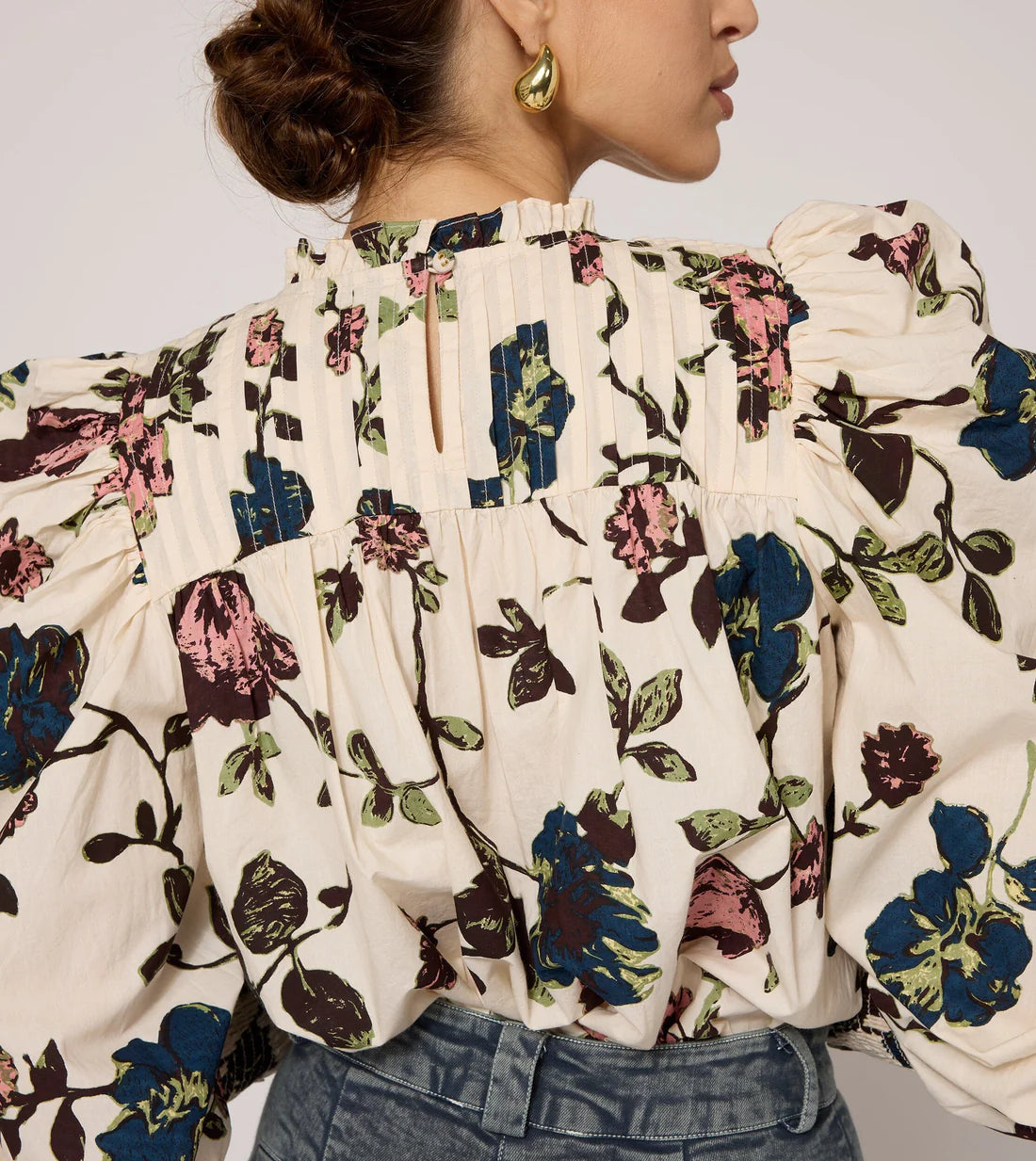 A woman is seen from the back, wearing the Irina Blouse by Cleobella, an ethically handmade, vintage-inspired top with a colorful floral pattern featuring blue and pink flowers and green leaves. The blouse boasts puffed sleeves and a high neck. Her hair is styled in a loose bun, complemented by gold hoop earrings.