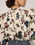 A woman is seen from the back, wearing the Irina Blouse by Cleobella, an ethically handmade, vintage-inspired top with a colorful floral pattern featuring blue and pink flowers and green leaves. The blouse boasts puffed sleeves and a high neck. Her hair is styled in a loose bun, complemented by gold hoop earrings.