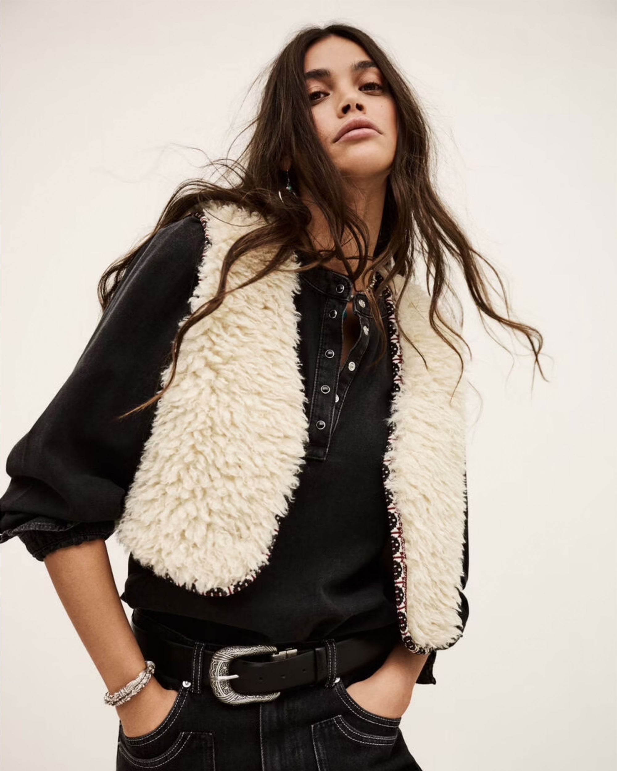 A woman with long, wavy dark hair wears the cream-colored BA&SH Jacket Celton over a black buttoned shirt and belted black pants made from washable fabric, posing confidently against a plain light background.