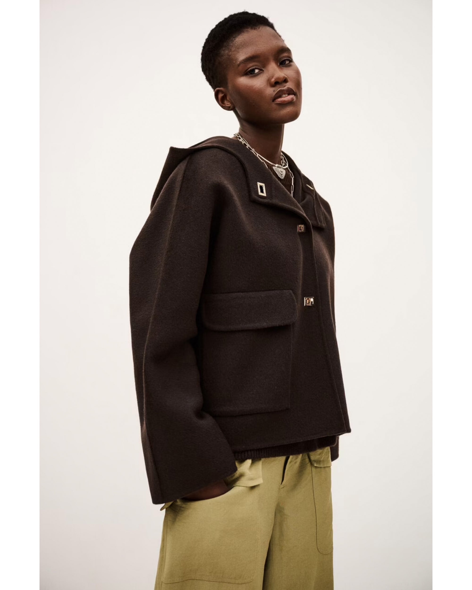 A person with short hair wears the BA&SH Jacket Kove—a loose, dark hooded jacket with large front pockets—paired with beige wide-leg pants, standing against a plain light background.