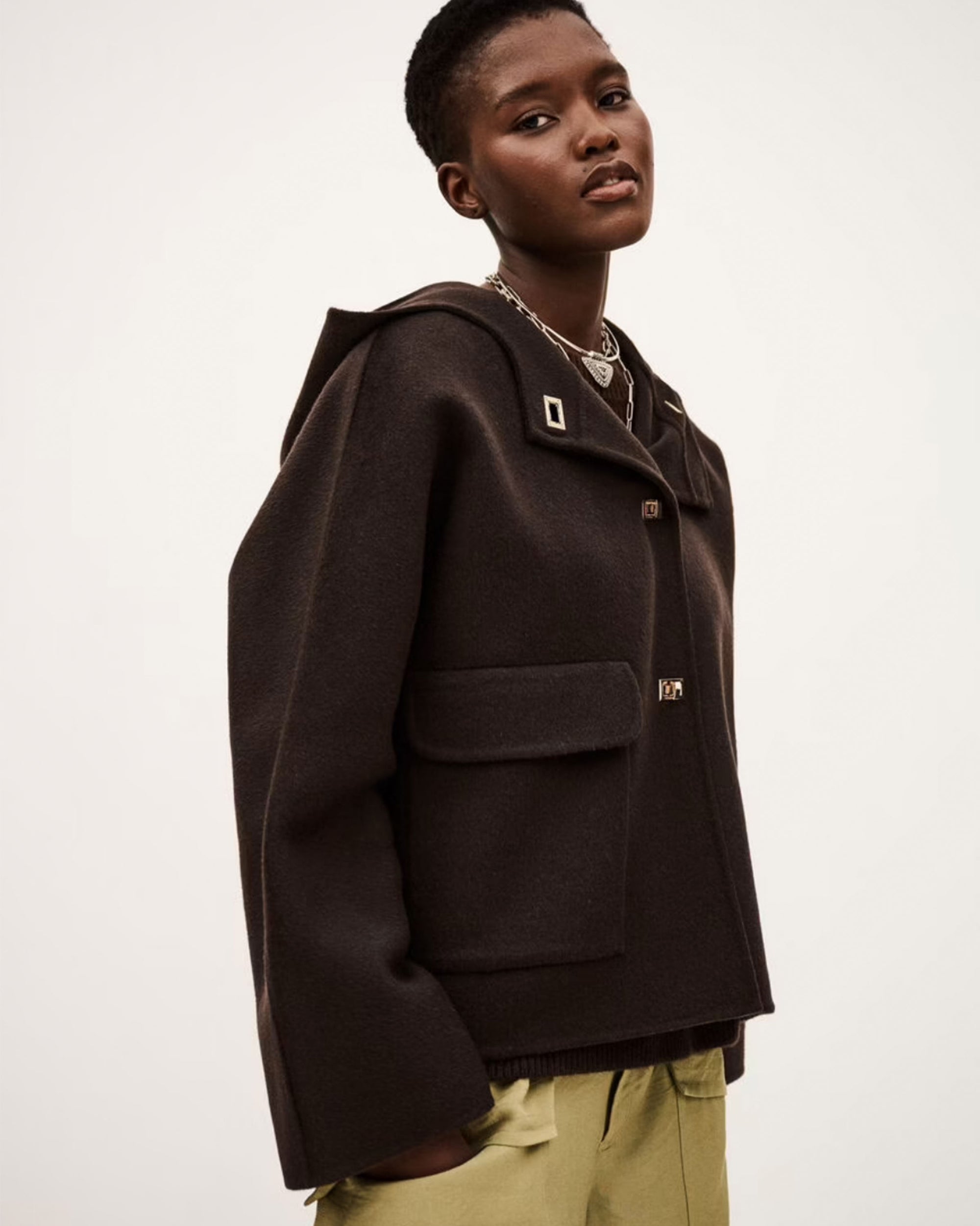 A person with short hair wears the loose-fitting, dark BA&SH Jacket Kove with large pockets and decorative buttons, paired with light green pants, standing against a plain light background.