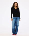 A woman with long, curly hair wears the Kerri Rosenthal Jackie Cardigan in black featherweight cashmere, styled with loose blue jeans, red pointed shoes, gold layered necklaces, and rings against a plain white background.