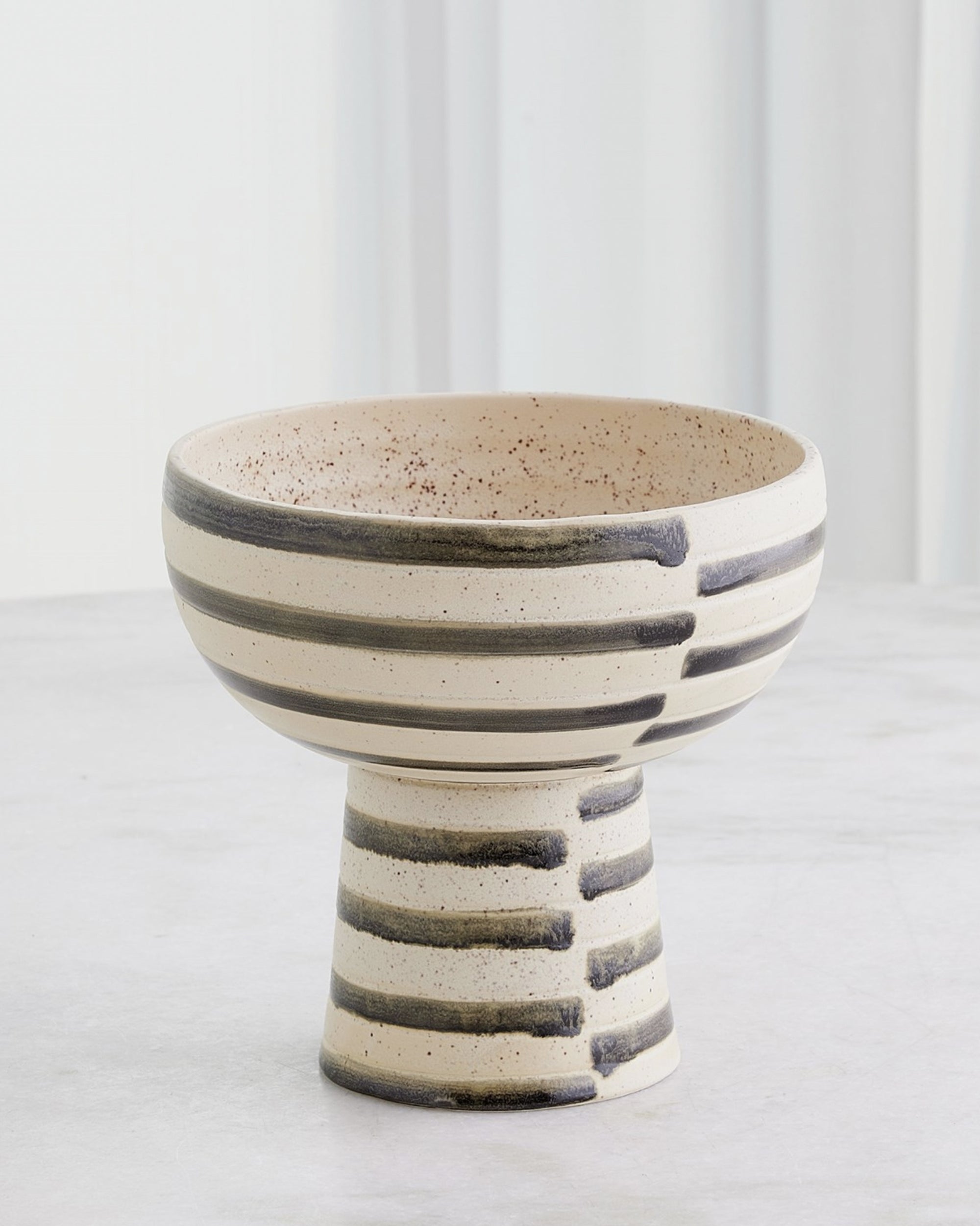 The 7.10640 Jail Break Footed Bowl by Global Views is a Portuguese ceramic piece with a wide, shallow top, tall narrow base, and hand-painted black stripes on off-white glaze. It’s displayed on a light surface with a blurred white background.