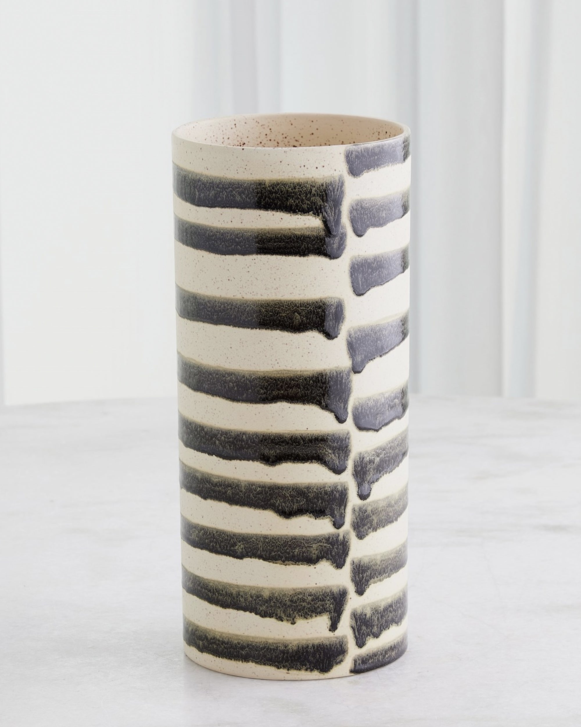 The 7.10641 Jail Break Cylindrical Vase L by Global Views is a tall Portuguese ceramic vase with a cream base and hand-painted black stripes, displayed on a light surface before white curtains; from the striking Jail Break Vases collection.