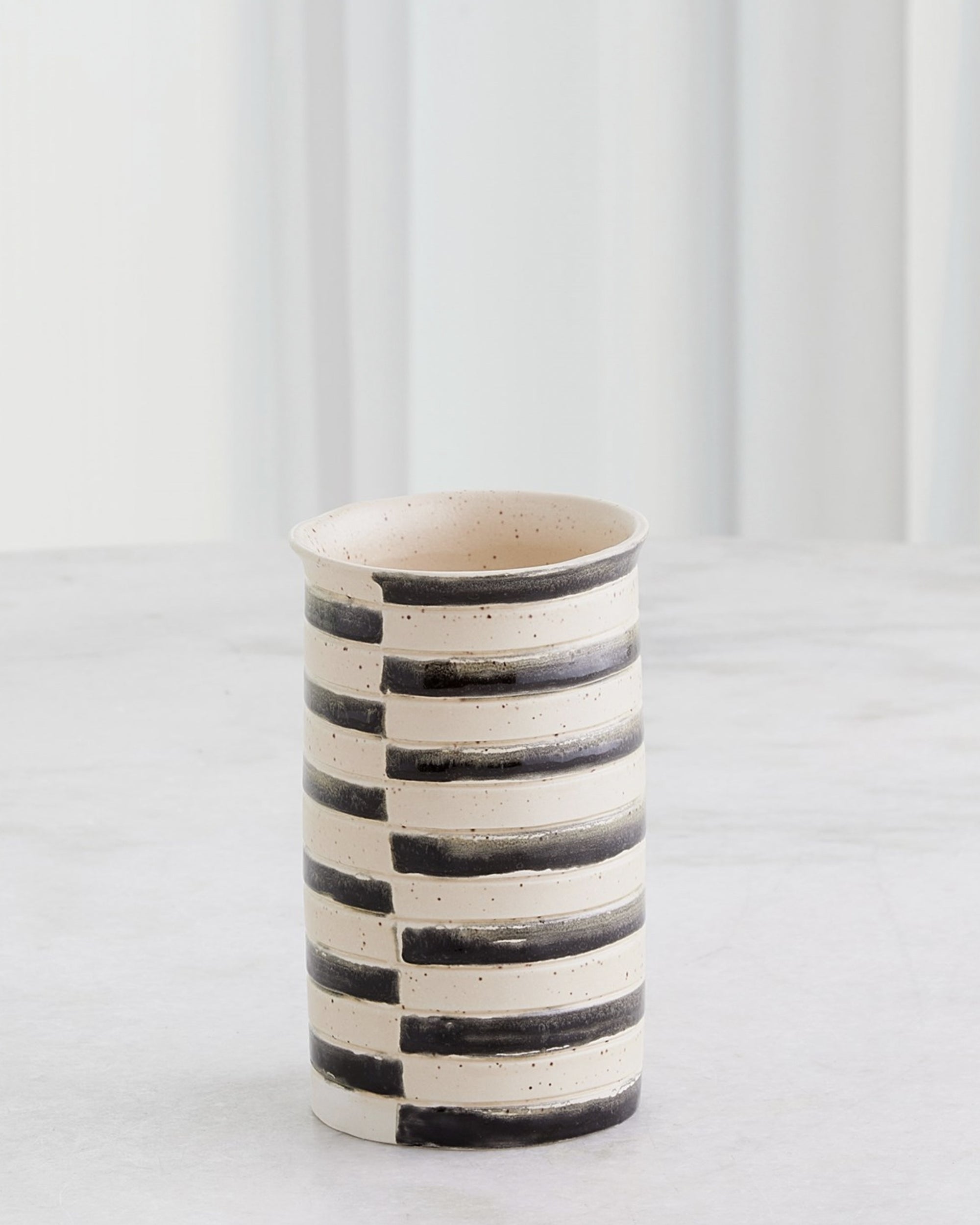 The Global Views 7.10642 Jail Break Cylindrical Vase S is a Portuguese ceramic vase with a cream base and hand-painted black stripes, shown on a light surface against a softly textured white background.